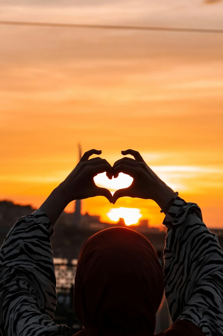Silhouette of a woman in a hijab making a heart shape with hands against a vibrant sunset.