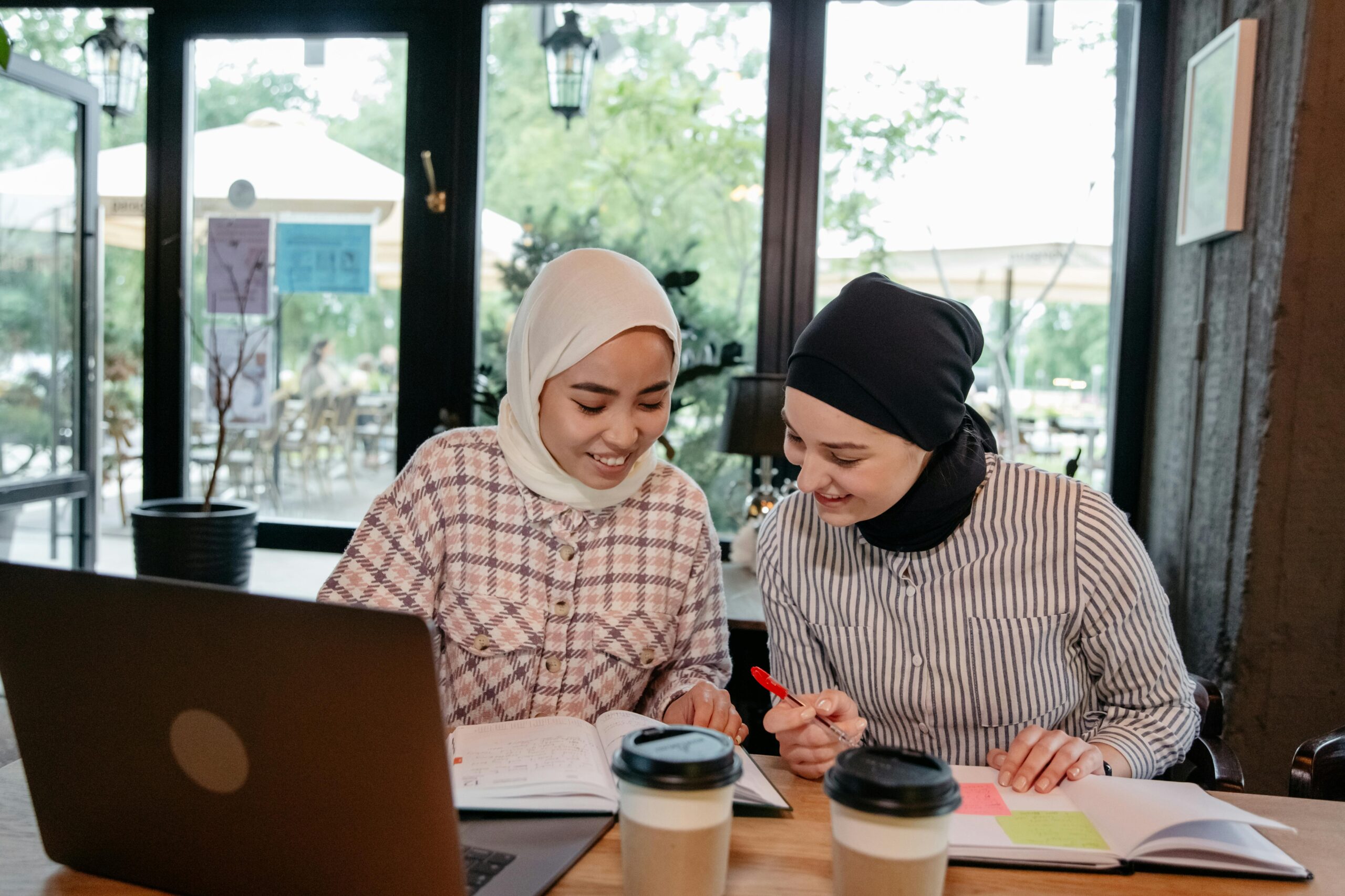 Two Muslim women in hijabs studying together with laptops in a cozy café.